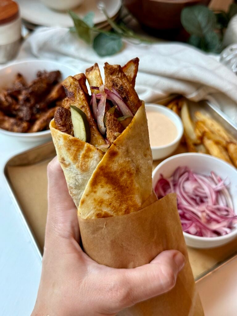 A hand holds a wrapped gyro filled with fries, grilled meat, pickles, onions, and creamy sauce. In the background are bowls of meat, red onions, sauce, and a tray of fries.