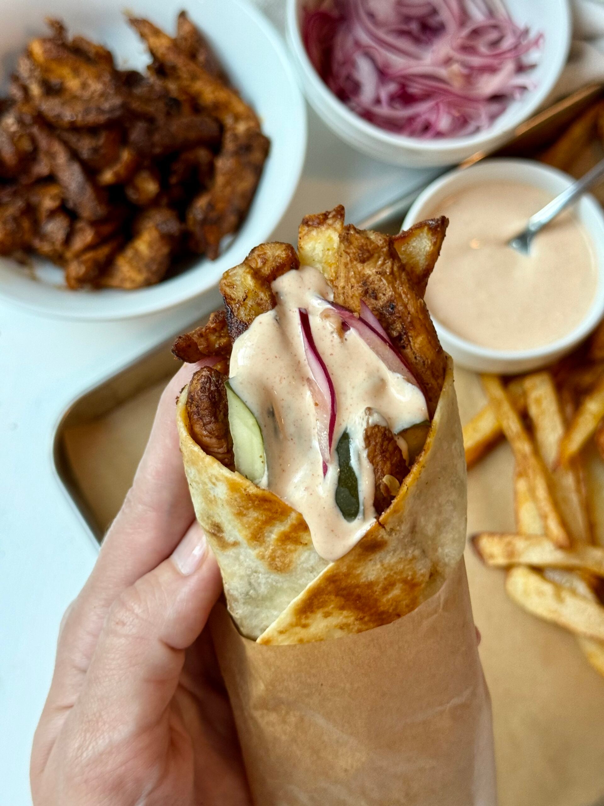 A hand holds a wrapped gyro filled with fries, grilled meat, pickles, onions, and creamy sauce. In the background are bowls of meat, red onions, sauce, and a tray of fries.