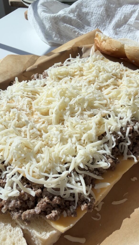 Open sandwich on brown parchment paper topped with ground beef, cheddar cheese slices, and a generous layer of shredded mozzarella cheese, ready for baking. The sandwich bun tops are visible in the background.