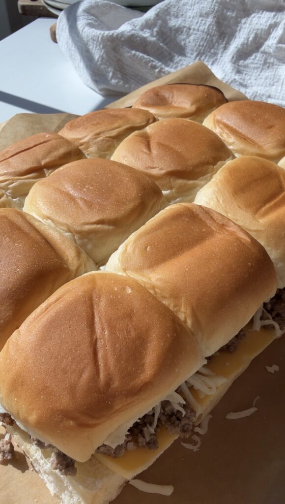 A close-up of several pull-apart slider sandwiches with golden brown tops, filled with cheese and ground beef on a baking tray.