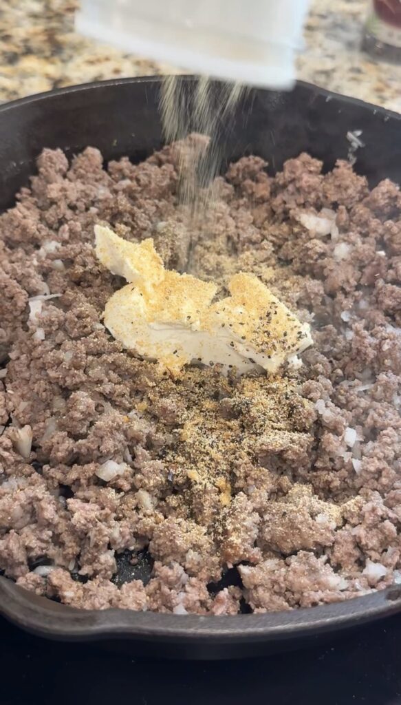 Ground beef and chopped onions cooking in a pan, with seasonings and a block of cream cheese added to the center.