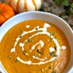 A bowl of creamy pumpkin soup garnished with a swirl of cream and sprinkled with pumpkin seeds, with small decorative pumpkins in the background.
