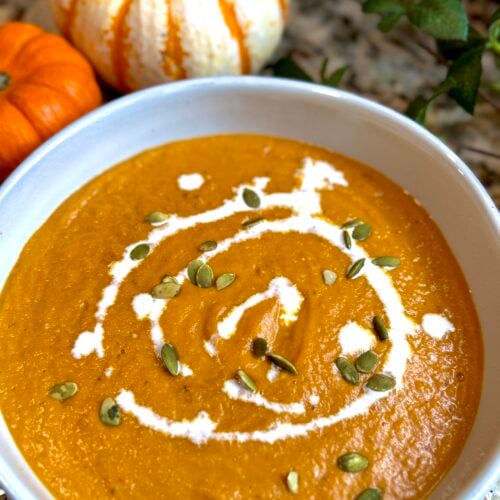 A bowl of creamy pumpkin soup garnished with a swirl of cream and sprinkled with pumpkin seeds, with small decorative pumpkins in the background.
