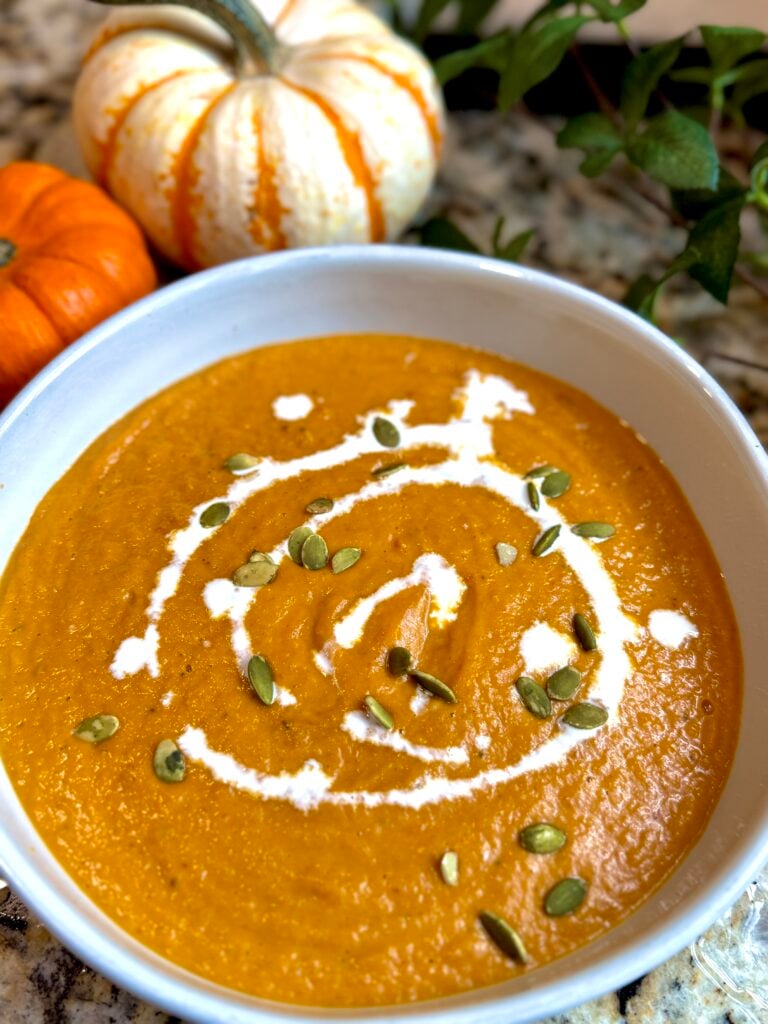 A bowl of creamy pumpkin soup garnished with a swirl of cream and sprinkled with pumpkin seeds, with small decorative pumpkins in the background.