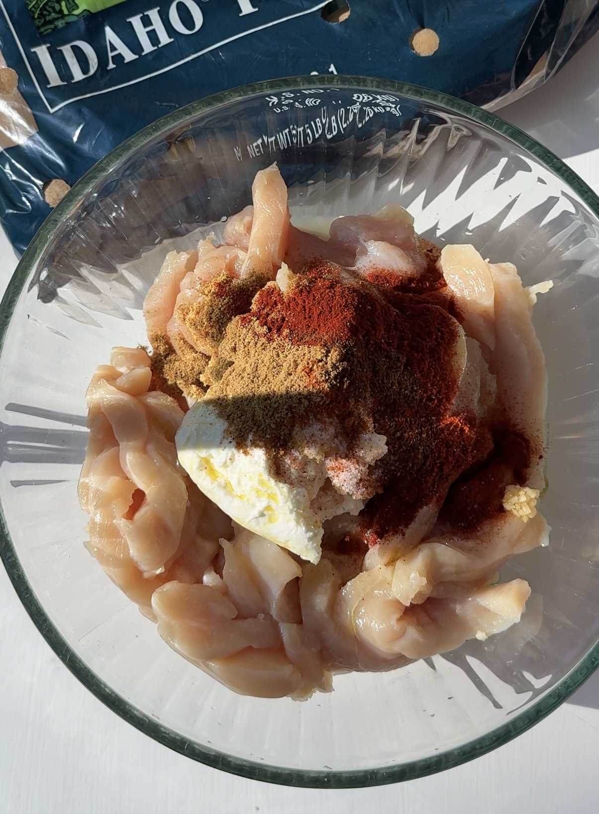 A glass bowl filled with raw chicken strips, yogurt, and various spices sits on a white surface. Part of a bag labeled IDAHO is visible in the background.