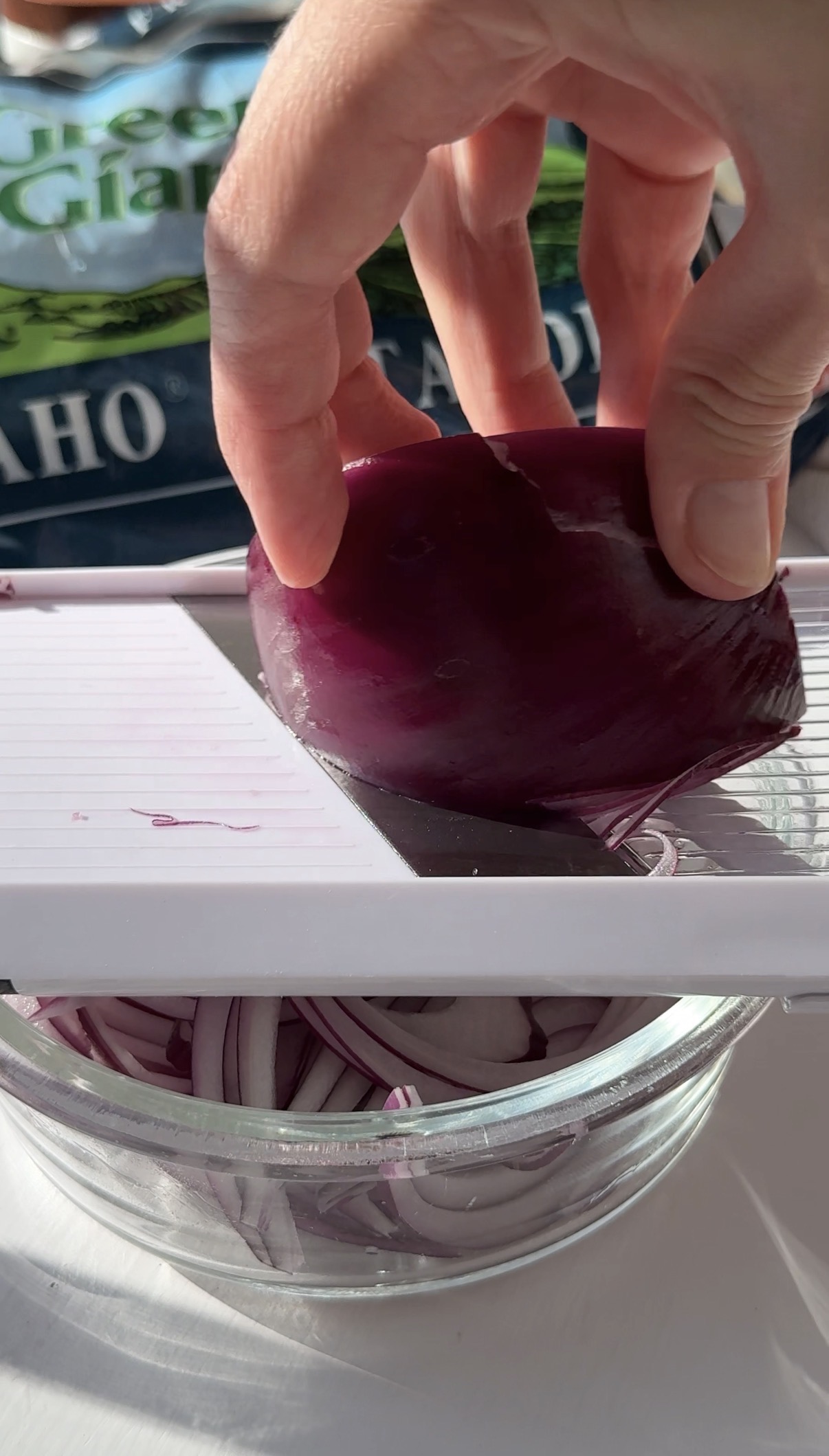 A hand slices a red onion on a white mandoline slicer over a glass bowl, with thin onion slices collecting below.