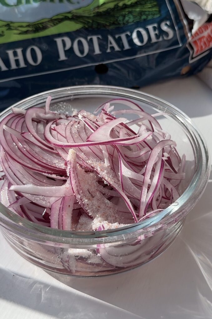 Thinly sliced red onions in a glass bowl sprinkled with salt. A bag of Idaho potatoes is partially visible in the background. Sunlight casts shadows across the scene.