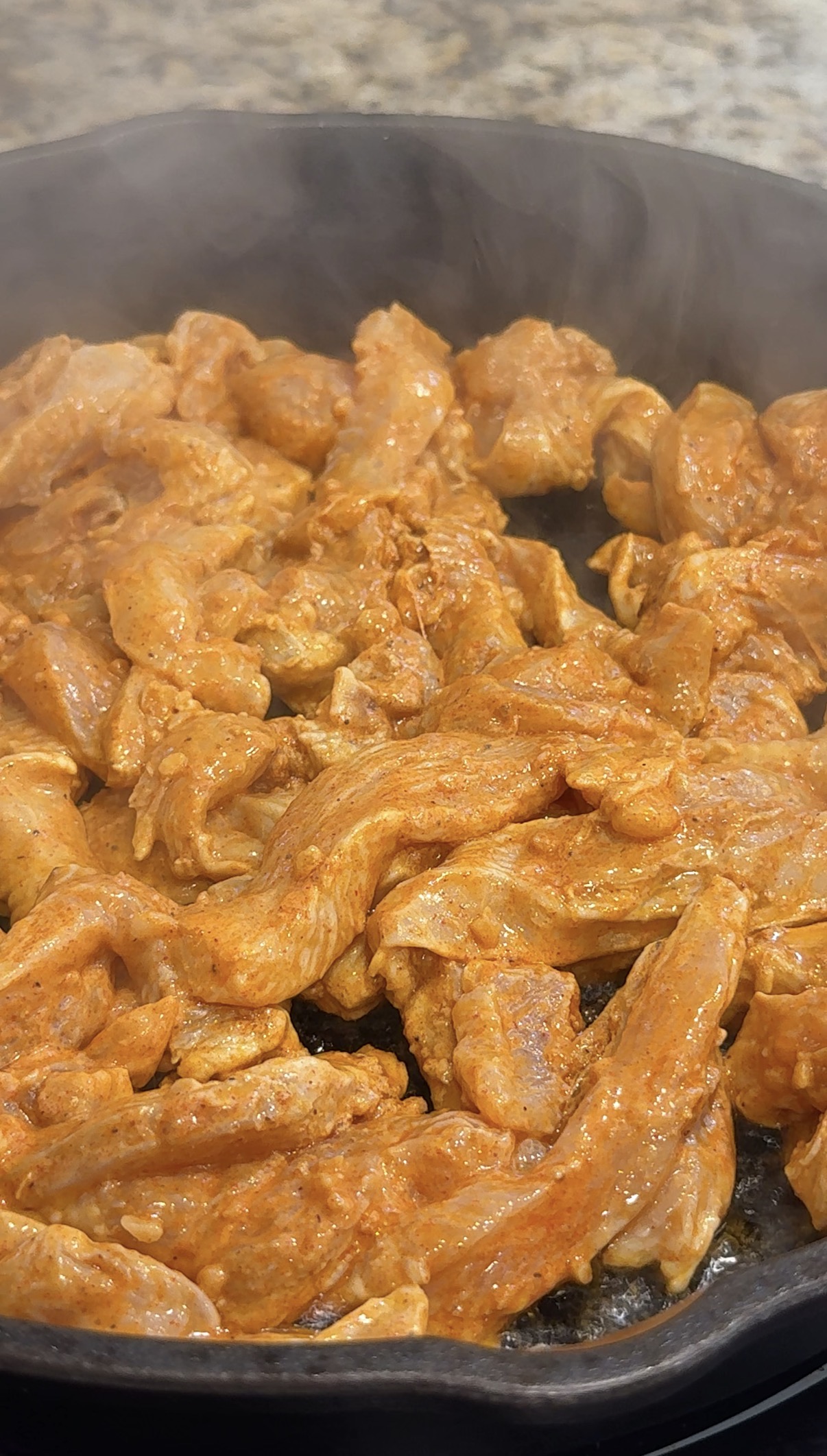 Strips of seasoned raw chicken cooking in a black skillet, with visible steam rising, on a speckled kitchen countertop background.