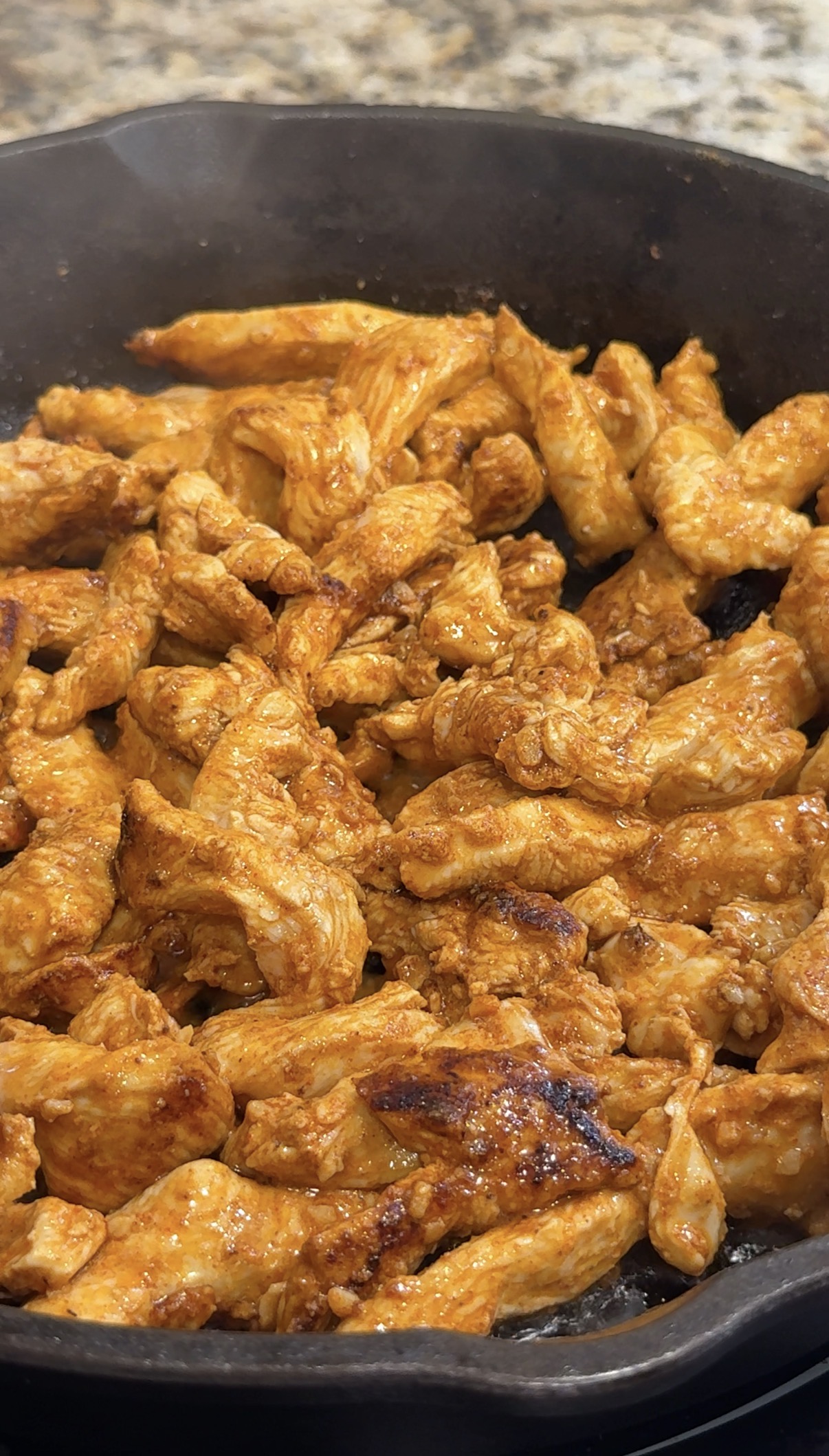 Cooked, seasoned chicken strips are shown in a pan. The chicken is browned and coated in a reddish-orange sauce, suggesting a savory, flavorful dish. The pieces appear tender and well-cooked, ready to be served or added to a recipe.