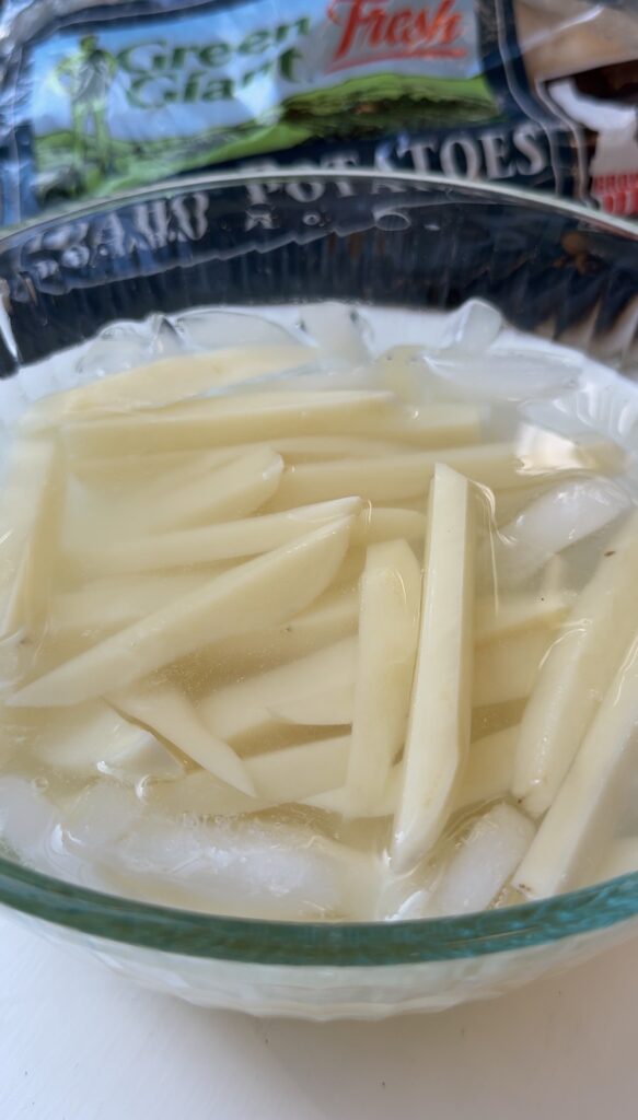 A glass bowl filled with water, ice, and raw potato slices soaking. The potatoes are cut into thin strips, resembling French fries.