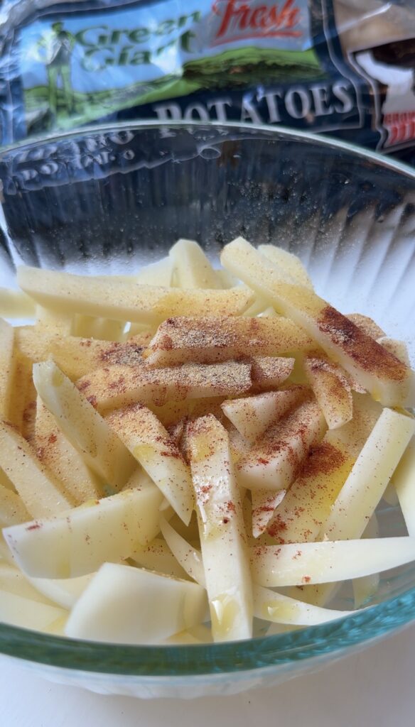 Raw potato sticks in a glass bowl are sprinkled with paprika and seasoning, ready for cooking. A potato bag is partially visible in the background.