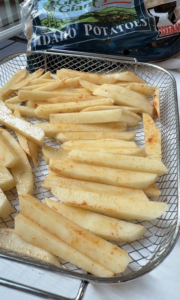 Raw potato fries seasoned with spices are arranged on a metal air fryer tray. In the background, there is a partially visible bag of Idaho potatoes.