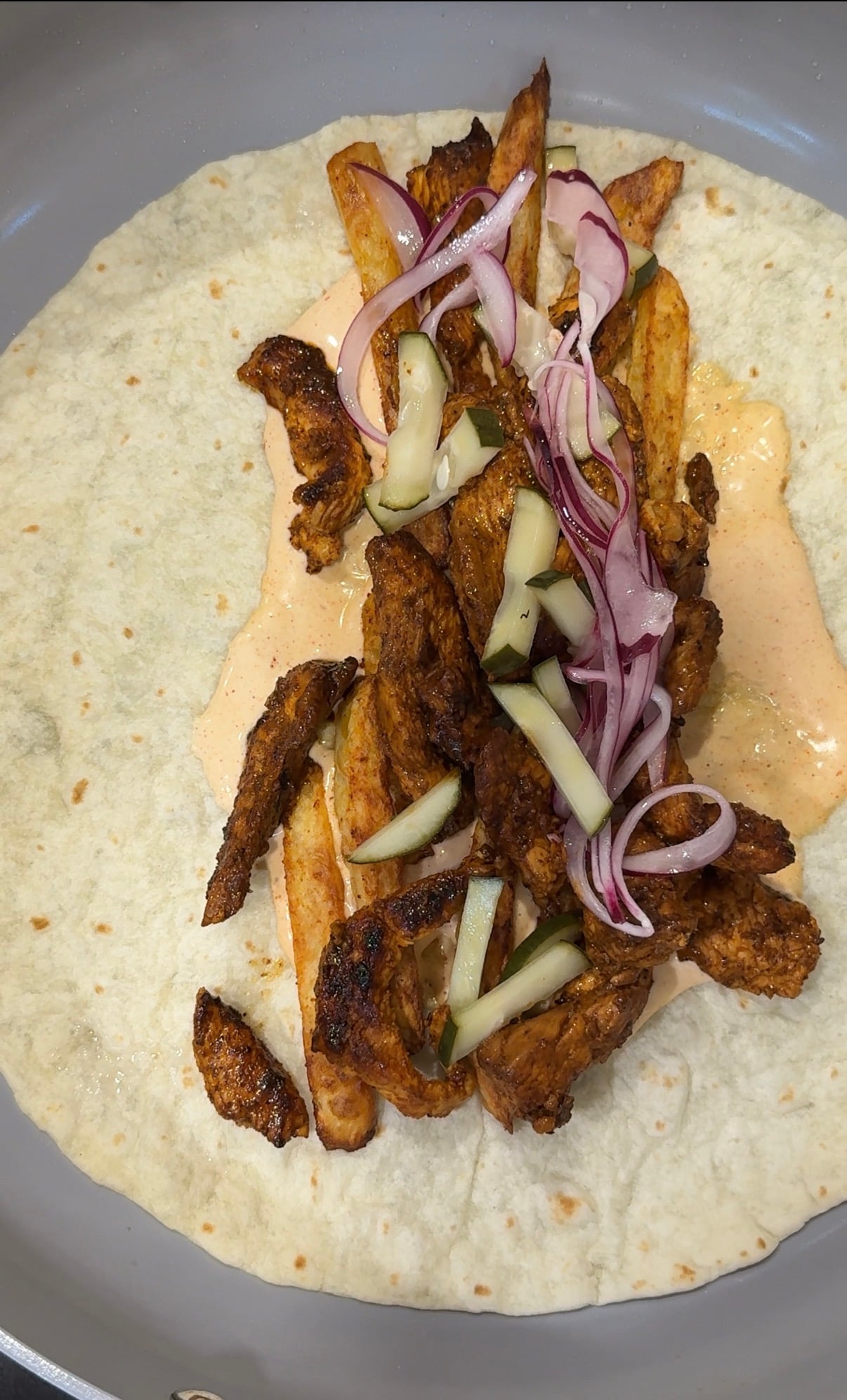 A tortilla topped with seasoned grilled chicken strips, sauce, thin cucumber slices, and red onion slivers, ready to be wrapped.