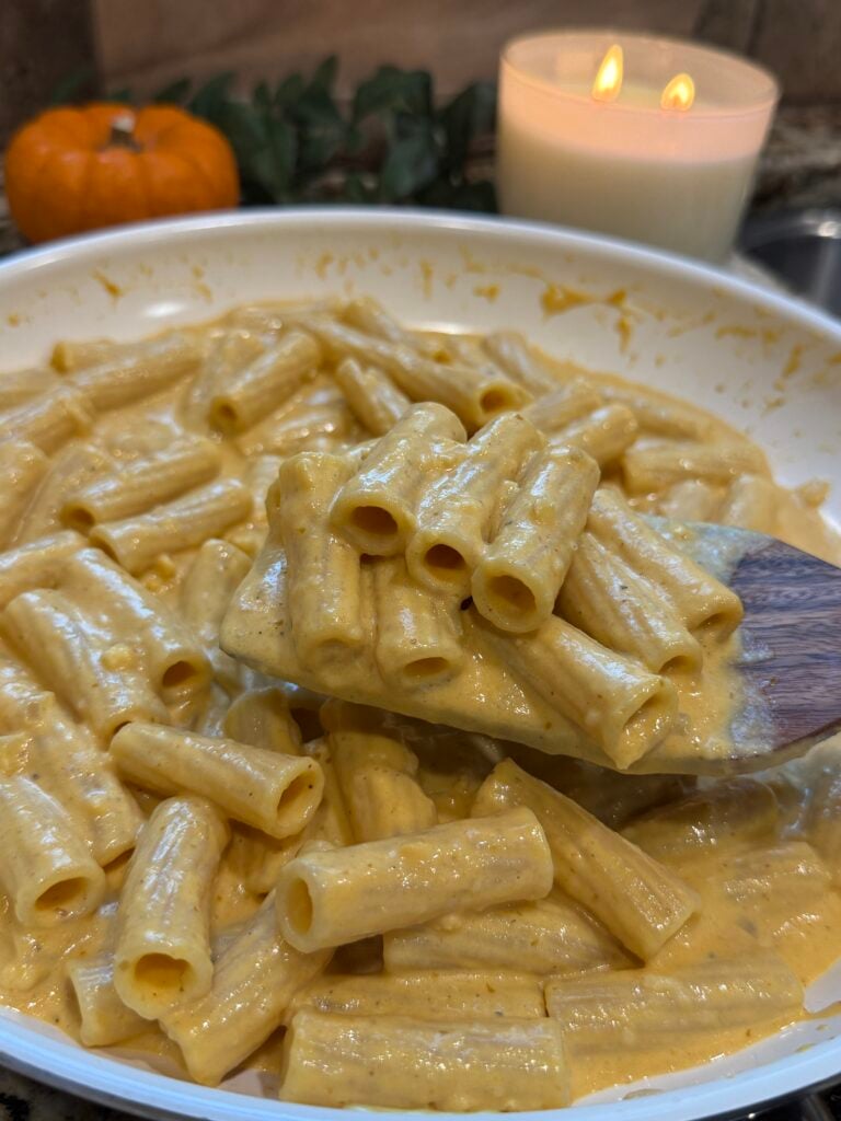 A wooden spoon holds creamy rigatoni pasta above a bowl. In the background, theres a lit candle, a small pumpkin, and green leaves.