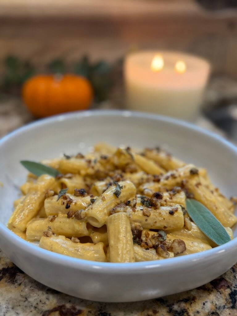A bowl of rigatoni pasta topped with a creamy sauce, crushed nuts, and fresh sage leaves. In the blurred background, there is a lit candle and a small orange pumpkin.