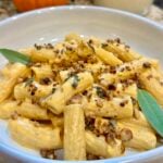 Creamy rigatoni pasta topped with chopped walnuts, fresh sage leaves, and a rich pumpkin-colored sauce, served in a white bowl.