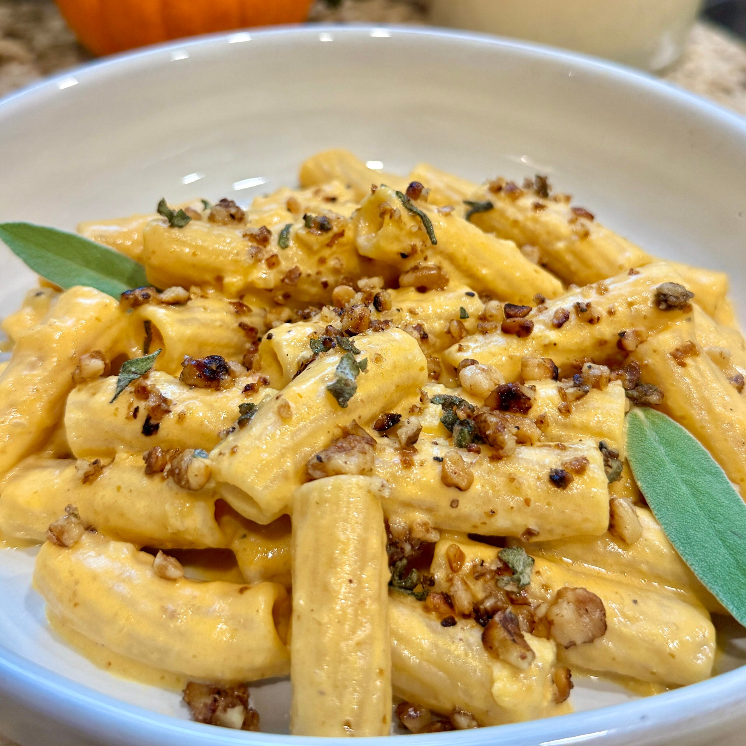 Creamy rigatoni pasta topped with chopped walnuts, fresh sage leaves, and a rich pumpkin-colored sauce, served in a white bowl.