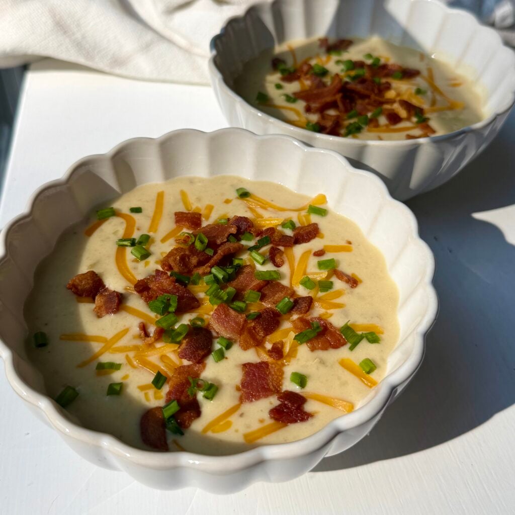 Loaded baked potato soup in two ceramic bowls topped with cheddar cheese, crispy bacon, sour cream, and green onions