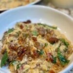 A close-up of a creamy pasta dish with orzo, sausage, sun-dried tomatoes, spinach, and melted cheese, served in a white bowl.