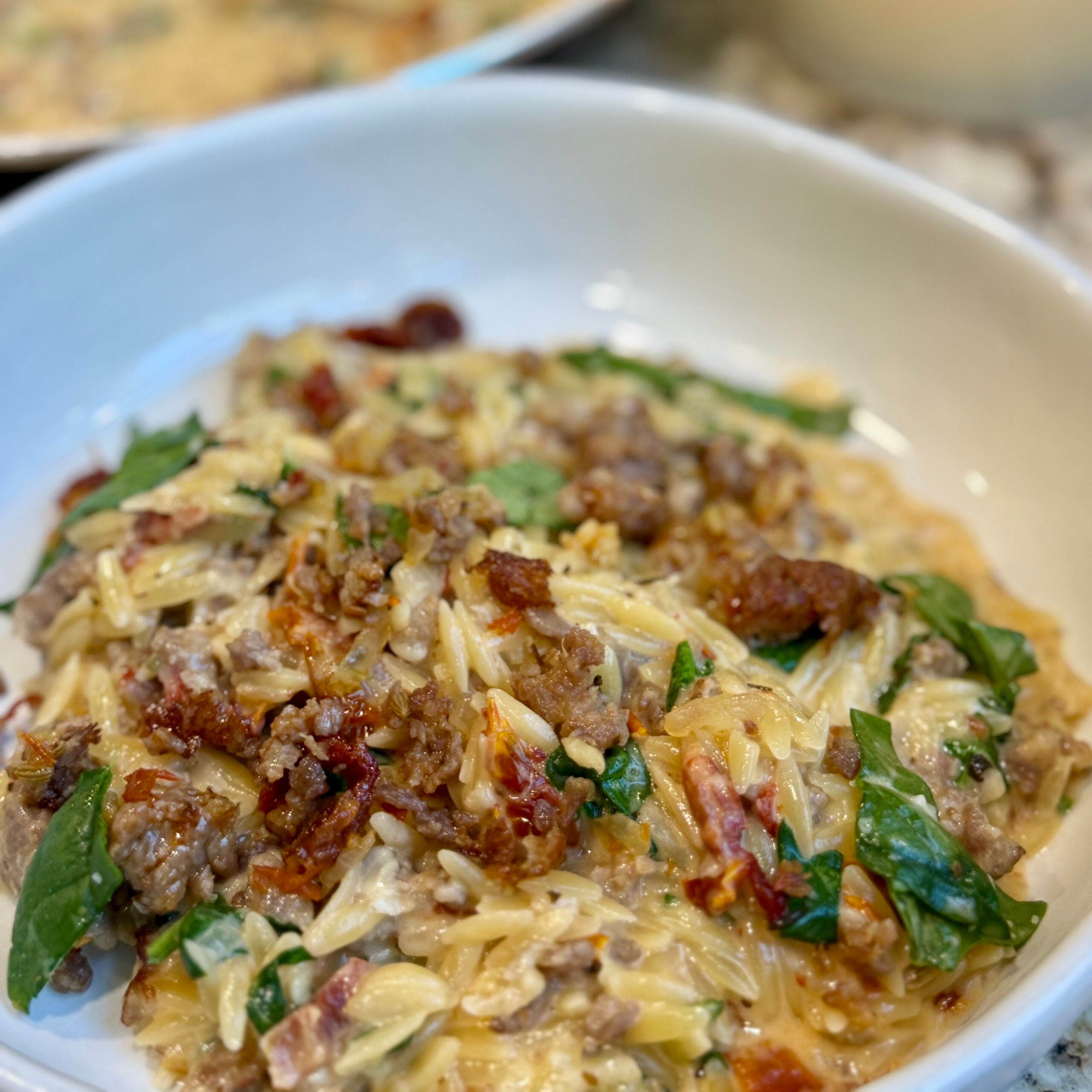 A close-up of a creamy pasta dish with orzo, sausage, sun-dried tomatoes, spinach, and melted cheese, served in a white bowl.