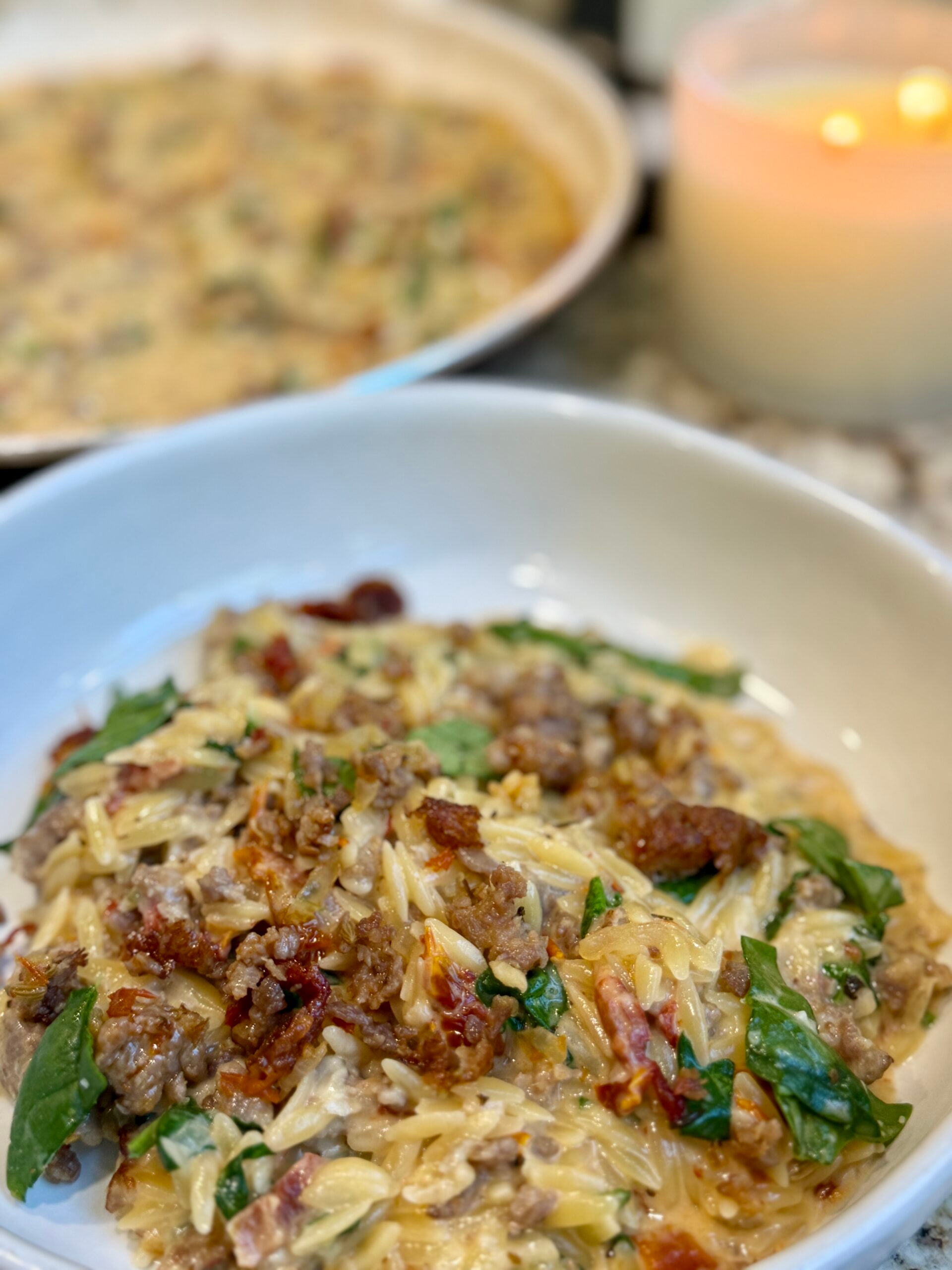 A close-up of a creamy pasta dish with orzo, sausage, sun-dried tomatoes, spinach, and melted cheese, served in a white bowl.