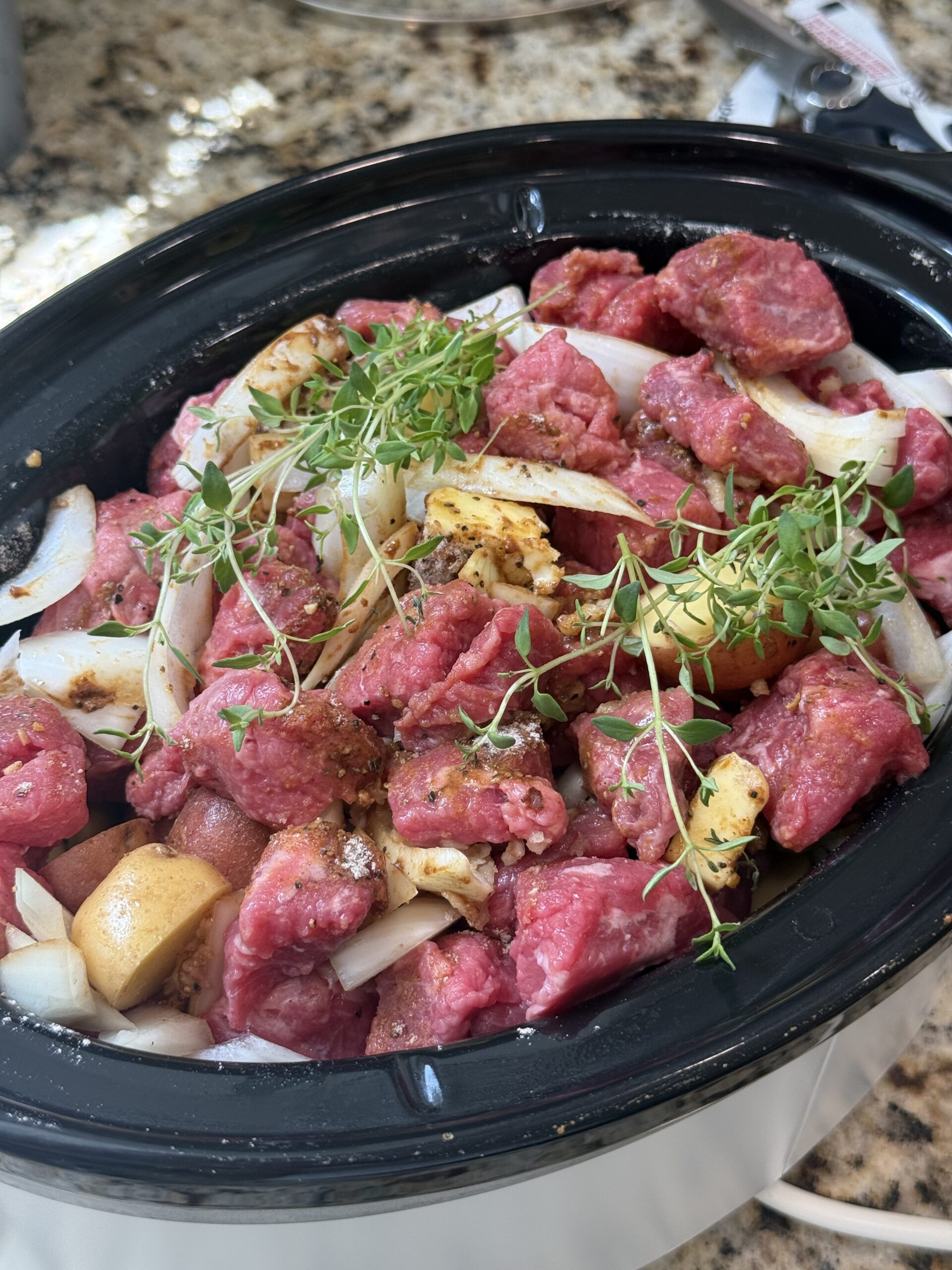 Raw steak bites and halved baby potatoes with garlic, onion, butter, thyme, and beef seasoning in a slow cooker